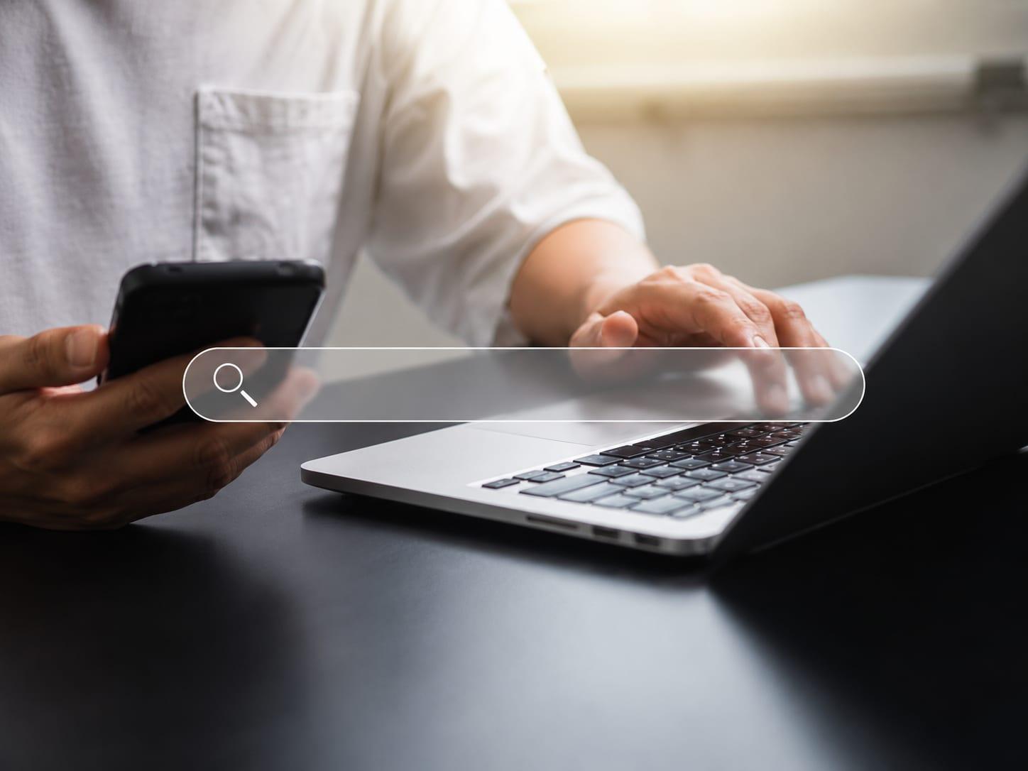 Person am Laptop mit Smartphone und eingeblendeter Suchleiste